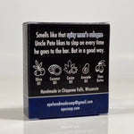 Back side of the For Pete's Sake soap. Quote on box says, "Smells like that spicy men's cologne Uncle Pete likes to slap on every time he goes to the bar. But in a good way." Box is black with dark blue accents.