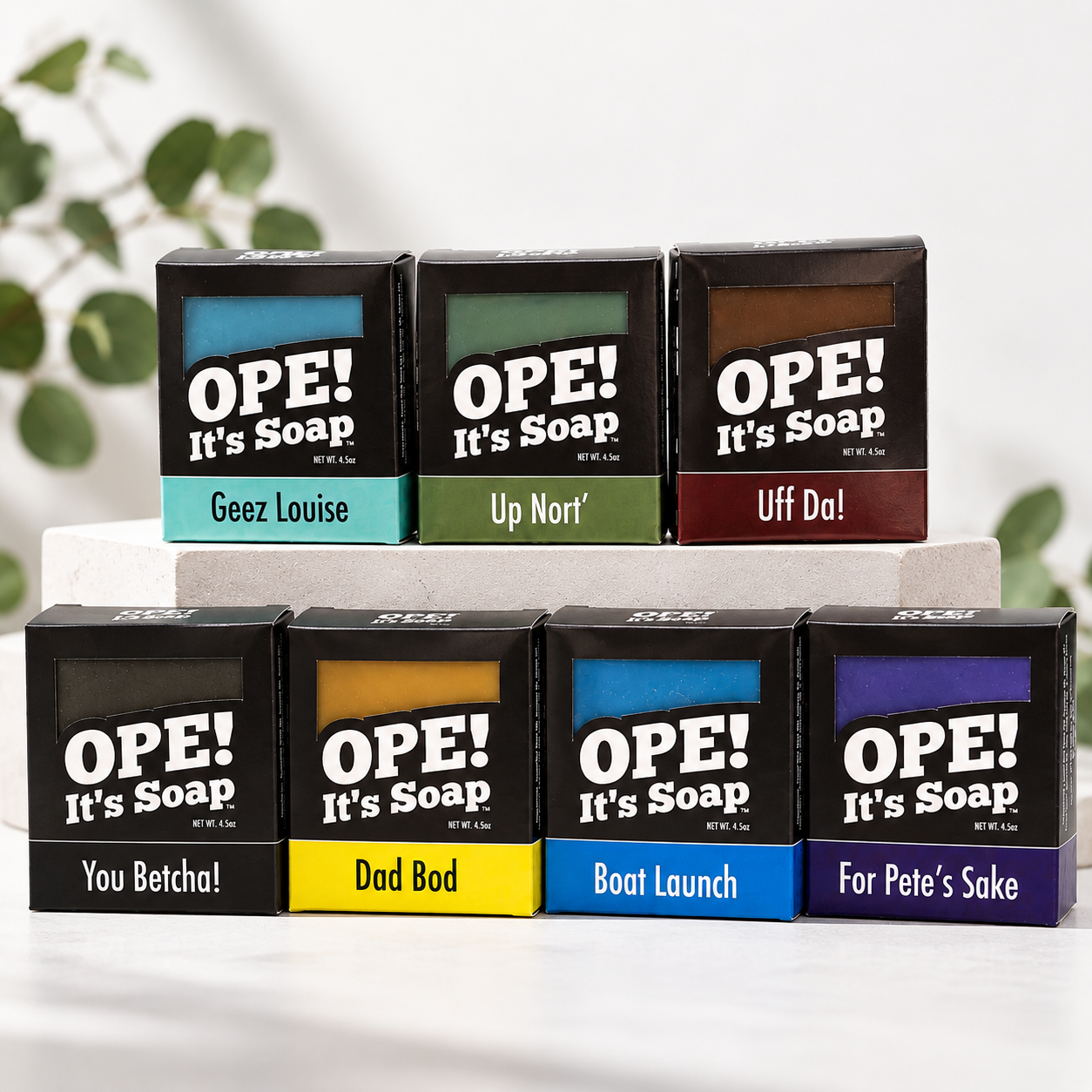 Seven Ope! It’s Soap Signature Collection bar soaps in colorful black packaging, arranged on a clean white display with soft greenery in the background.