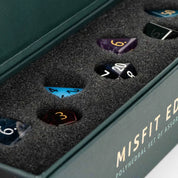 Open dark green presentation box containing a Misfit Edition polyhedral set of assorted dice nestled in black foam. Visible dice include blue, purple, black, and brown polyhedra with white, gold, and red numbering.