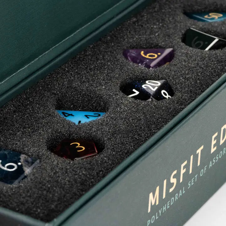 Open dark green presentation box containing a Misfit Edition polyhedral set of assorted dice nestled in black foam. Visible dice include blue, purple, black, and brown polyhedra with white, gold, and red numbering.