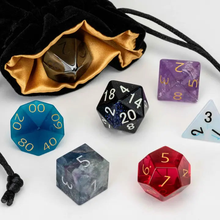 A set of translucent polyhedral gaming dice with gold numbering, displayed alongside a black velvet drawstring bag with a gold interior on a white background.