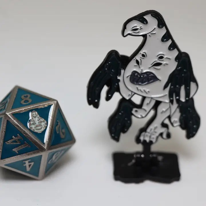 A black and silver gargoyle enamel pin standing on a small black base, displayed next to a blue and silver 20-sided die on a plain light background.