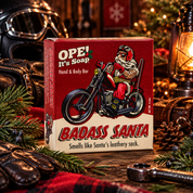 Ope! It’s Soap Badass Santa soap box displayed in a rugged holiday scene with leather motorcycle gear, evergreen branches, pinecones, a glowing lantern, and warm Christmas lights.