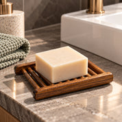 A rectangular bar of cream soap resting on a dark brown bamboo soap dish, placed on a gray stone bathroom countertop next to a folded green towel and a white sink with a gold faucet.