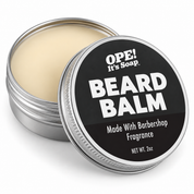 An open round metal tin of OPE! It's Soap Beard Balm, showing the light beige balm inside, with the lid leaning in front displaying a black label with white text.