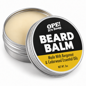An open round metal tin of OPE! It's Soap Beard Balm, showing the pale cream balm inside, with the lid leaning in front. The lid features a black and yellow label with bold white text.