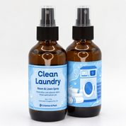 Two Clean Laundry Room & Linen Spray bottles with light blue labels, featuring a washing machine and folded towels, handmade by Coleman & Peck in Chippewa Falls, Wisconsin.