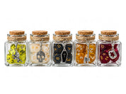 Five clear glass dice jars with cork stoppers and twine accents, featuring colorful polyhedral gaming dice in yellow, white, black, amber, and burgundy. Each jar has a unique silver charm, including a dragon, skull, coffin, axe, and fangs, displayed on a white background.