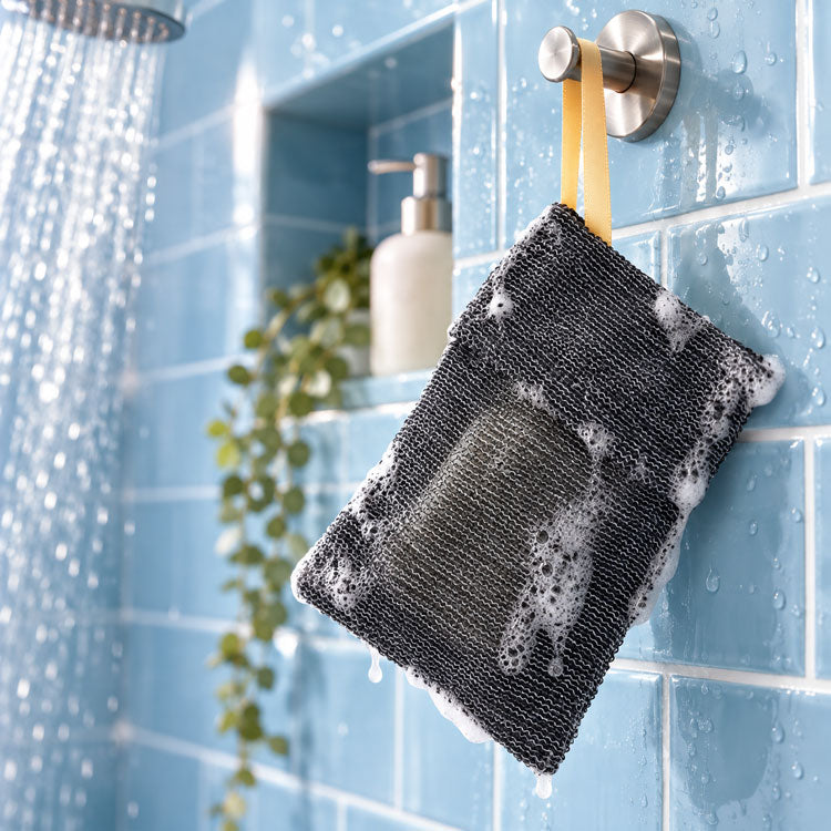 Black textured exfoliating soap saver bag with a bar of soap inside, covered in white suds and hanging by a gold loop from a brushed metal hook in a modern shower with light blue subway tiles, running water, and a recessed shelf with a plant and soap dispenser.