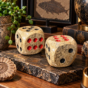 Two decorative fossilized coral dice with natural tan stone patterns and black and red dots displayed on a dark stone slab, surrounded by fossils, books, and mineral specimens in a warmly lit collector’s study.