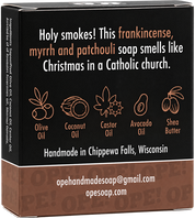 Back of the Ope! It’s Soap Holy Smokes soap box featuring the tagline “Holy smokes! This frankincense, myrrh and patchouli soap smells like Christmas in a Catholic church,” with illustrated icons highlighting the soap’s plant oils and shea butter.