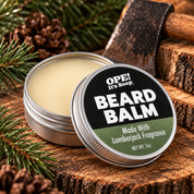 An open round tin of Ope! It's Soap Beard Balm sits on a rustic wood surface with pine branches, pinecones, and a wooden-handled tool in the background. The lid is placed in front of the tin, showing the label text 'BEARD BALM' and 'Made With Lumberjack Fragrance'.
