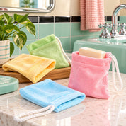 Four reusable microfiber soap bags in pink, blue, yellow, and green, shown hanging on and resting on a bathroom counter next to a sink.