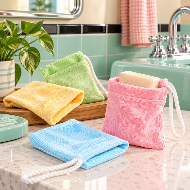 Four reusable microfiber soap bags in pink, blue, yellow, and green, shown hanging on and resting on a bathroom counter next to a sink.