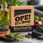 Ope! It’s Soap Natural Facial Bar displayed in a modern botanical scene with tea tree leaves, golden oil, tamanu nuts, activated charcoal, and the black charcoal soap visible through the box window.