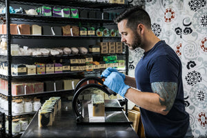 Image of Tony, the soap maker and owner of Ope! It's Soap, focused while cutting a batch of soap in his soap studio. A shelf of soap and body butters is seen in the background.