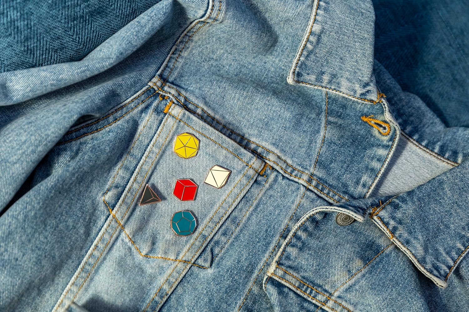 Five colorful Platonic Solids enamel pins representing tabletop gaming dice shapes D4, D6, D8, D12, and D20, displayed on the front pocket of a light blue denim jacket.