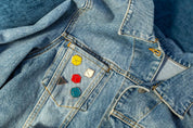 Five colorful Platonic Solids enamel pins representing tabletop gaming dice shapes D4, D6, D8, D12, and D20, displayed on the front pocket of a light blue denim jacket.