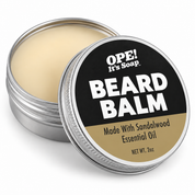 An open round metal tin of OPE! It's Soap Beard Balm, showing the pale cream balm inside, with the lid leaning in front. The lid features a black and tan label with bold white text.