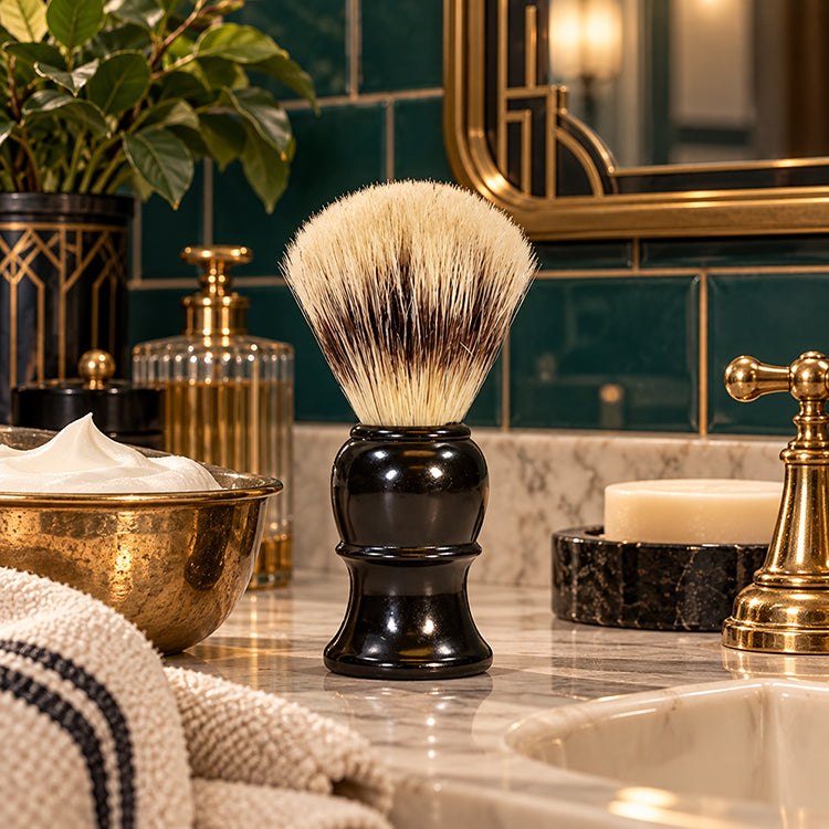 A shaving brush with dense cream and dark brown bristles on a glossy black handle, displayed on a marble bathroom counter with a gold faucet, soap dish, and green tiled wall in the background.