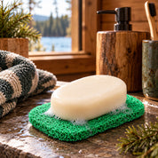 Green textured soap saver pad holding a white bar of soap with light suds, resting on a wet stone countertop in a modern cabin bathroom with wood accents, folded towels, and a window overlooking a lake and pine trees.