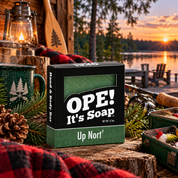 Ope! It’s Soap Up Nort’ soap box displayed in a cozy Wisconsin cabin scene with pine branches, fishing gear, a camp mug, lantern, plaid flannel, and a peaceful lake at sunset.