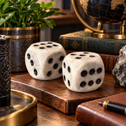 Two oversized white marble dice with black dots displayed on a wooden tray in a warmly lit office setting, surrounded by vintage books, a decorative globe, greenery, and natural stone accents.
