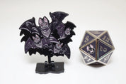 Black and purple die with bat design on a stand next to a metal polyhedral dice with similar design